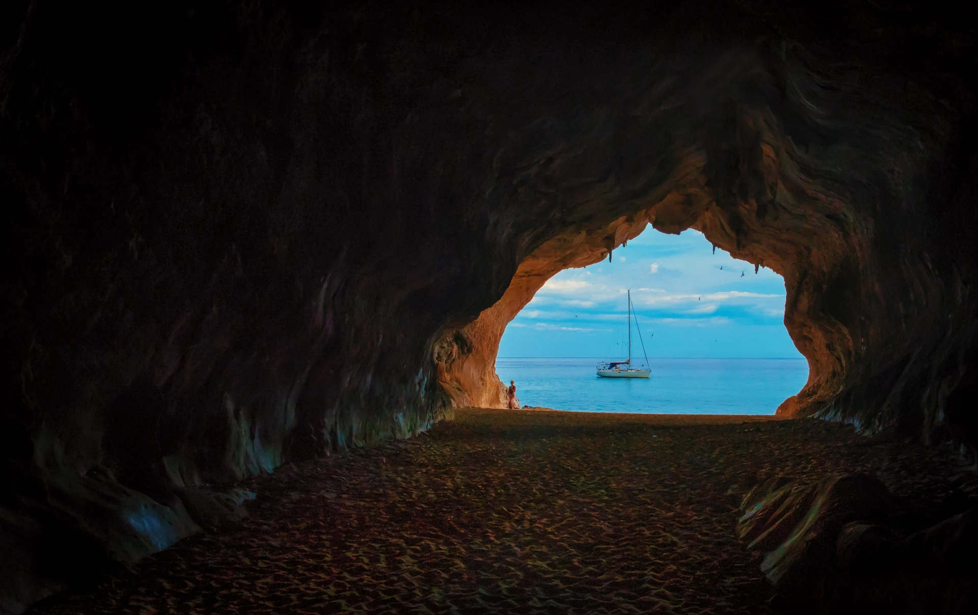 A sea cave opens onto blue water, with a sailboat beyond.